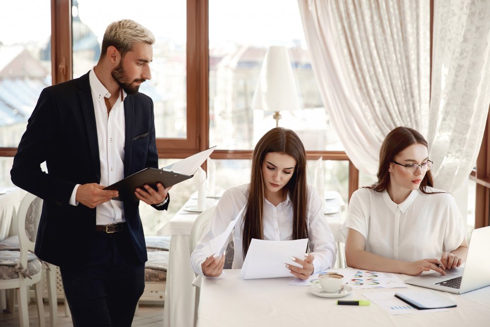 Business team discussing succession planning strategies in a modern office setting, with a man holding a folder and two women reviewing documents and a laptop.
