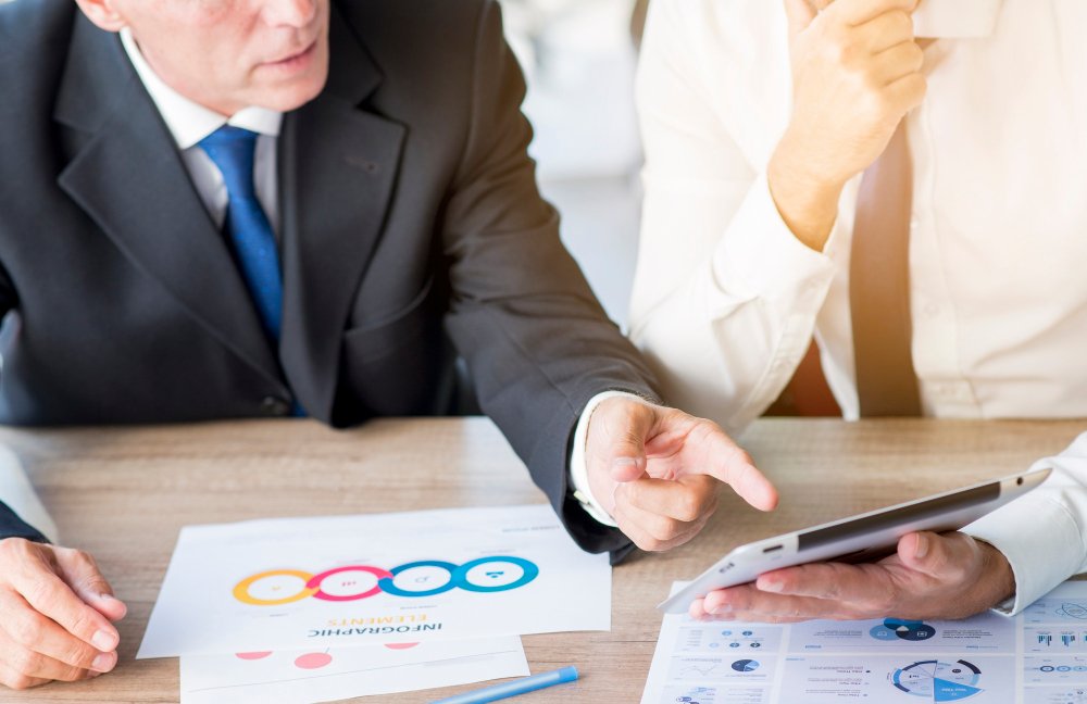 Business professionals discussing corporate governance strategies over a tablet, with documents and charts on a table.