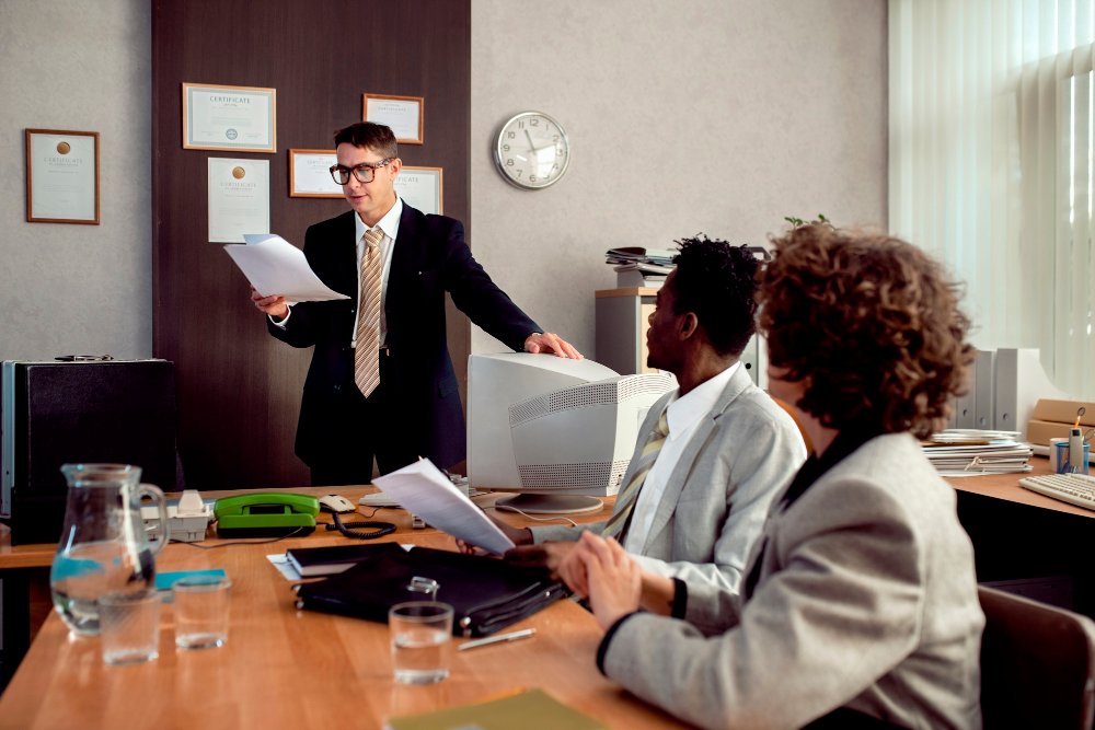 Business meeting with a lawyer presenting S-Corporation setup information to clients in an office setting, featuring legal documents and certificates on the wall.