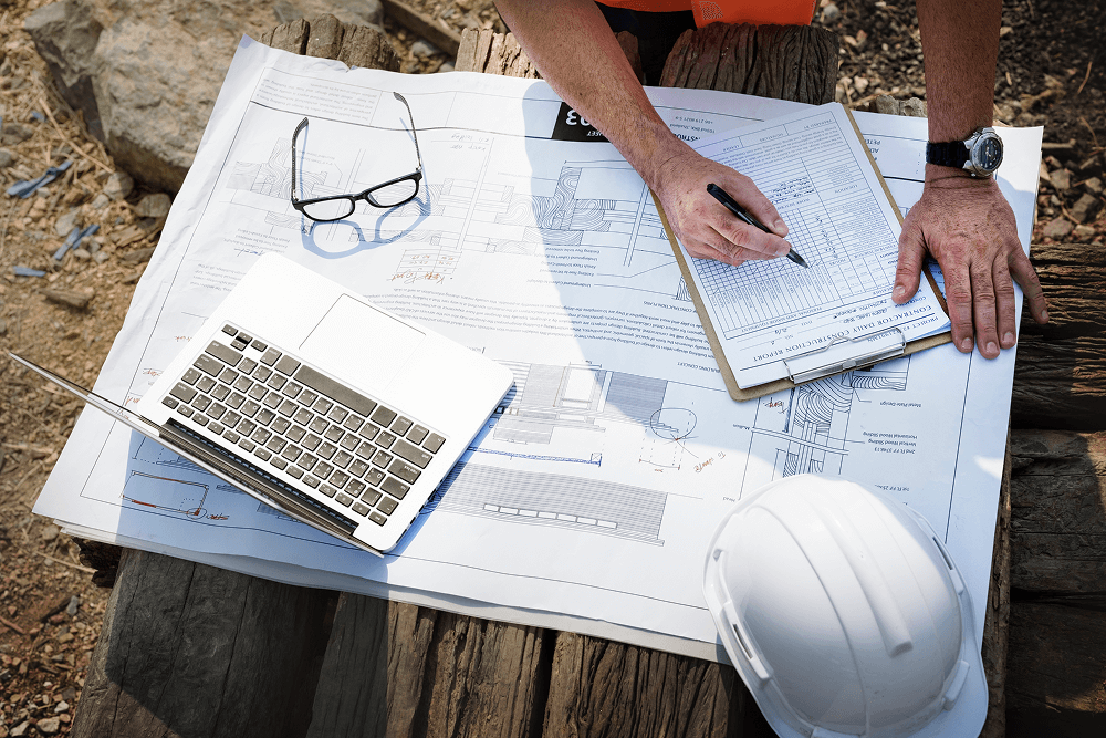 Construction professional reviewing blueprints and compliance documents on a job site, with a laptop and safety helmet nearby.