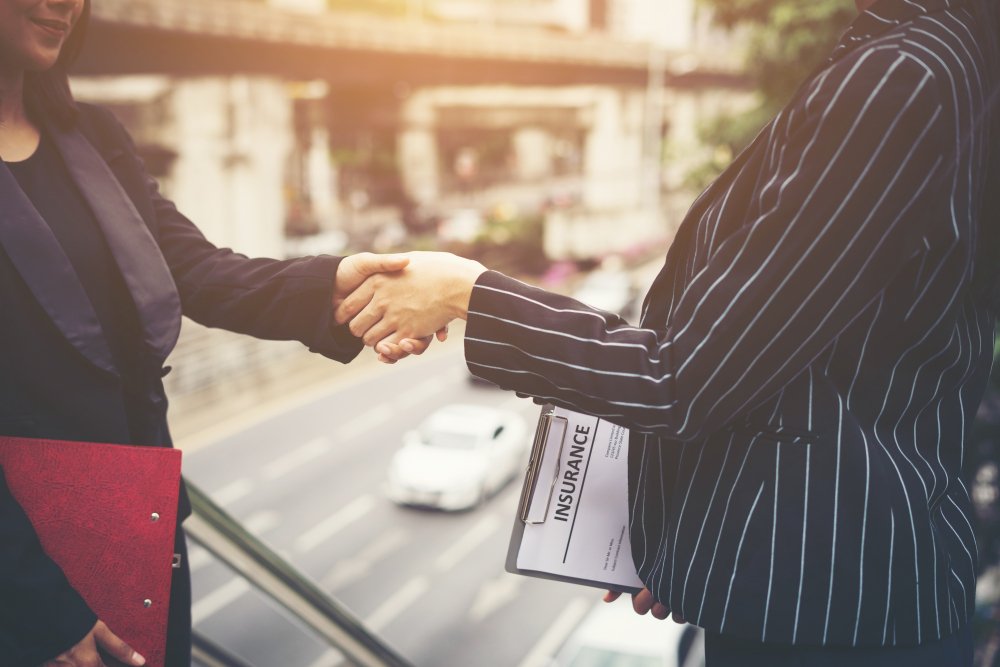 Business professionals shaking hands, symbolizing partnership and agreement in mergers and acquisitions, with a clipboard labeled "INSURANCE" visible, set against an urban backdrop.