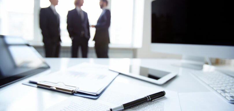Business professionals discussing in an office setting, with a clipboard, documents, and electronic devices in the foreground, reflecting business law consultations and contract discussions.