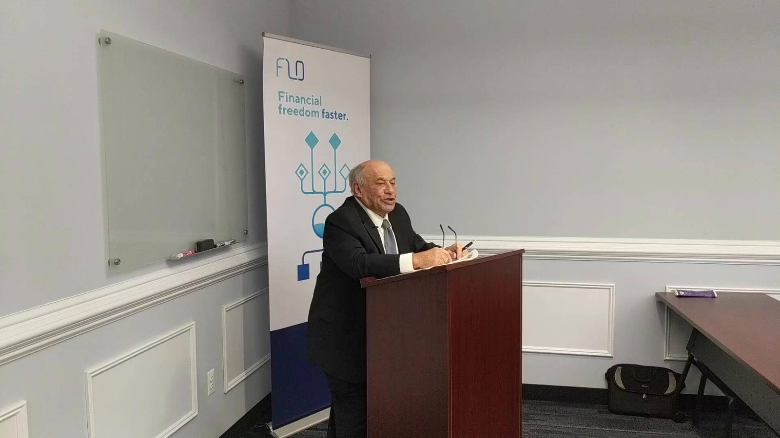 Elderly man speaking at podium with legal and financial branding, promoting business law services and financial strategies.