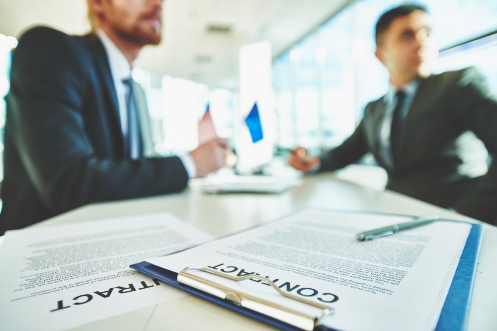 Business professionals discussing a contract during mediation session, with focus on document labeled "CONTRACT" and flags in background.