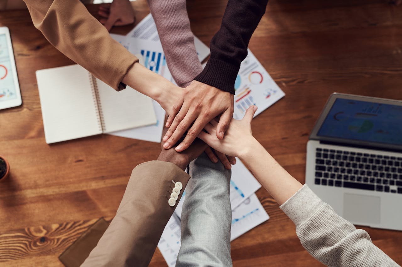Hands of diverse individuals joining in a collaborative gesture over a wooden table with business documents and a laptop, symbolizing teamwork and startup legal support.