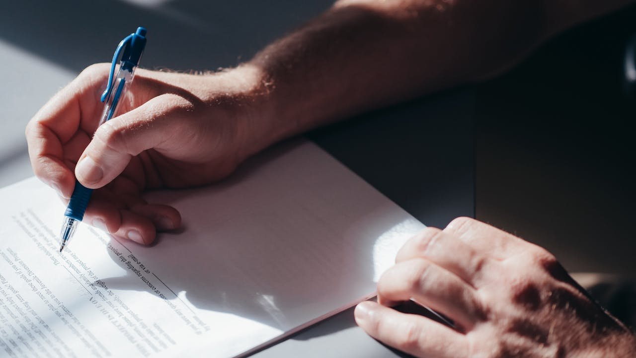 Hand holding a blue pen writing on a document, representing independent contractor agreements and legal services in New Jersey.